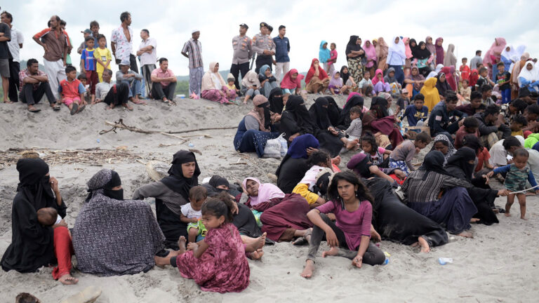 Dozens of men, women and children sit or stand on the beach, some laying down. Several Indonesian officials stand among them.