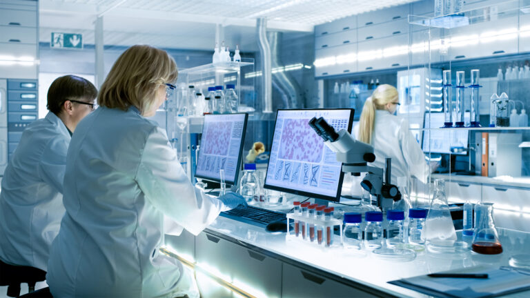 Female and male scientists working on Computers In a modern laboratory with various shelves, beakers, chemicals and other equipment visible.