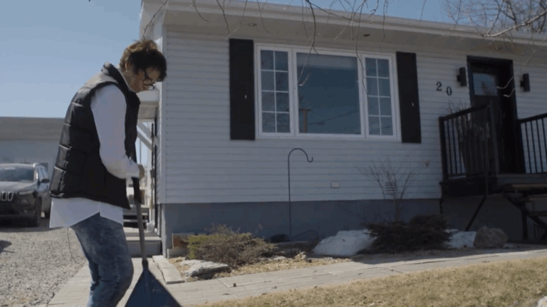 Wylde is sweeping a broom in front of the one-storey home. The home has a light-coloured siding, and the living-room window has a white frame and thin black shutters. Wylde is dressed in jeans, a white shirt and a black puffer vest. She has short brown hair and dark-rimmed glasses.