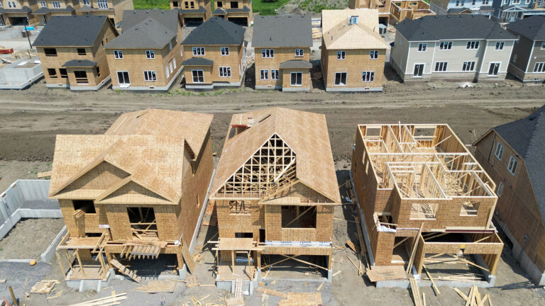 Dozens of partially built single-family two-storey homes sit tightly packed. Two of them have partial roofs built. Lumber is scattered around three of the houses, which are all clad in wood panels in the beginning stage of construction. A dirt road separates the homes in the foreground from seven homes on the other side of the street-to-be. In the distance is a green space.