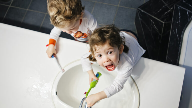 Seen from above, two white toddlers hold toothbrushes as they lean toward a bathroom sink, One of them has a hand on the faucet and is looking up with mouth open.
