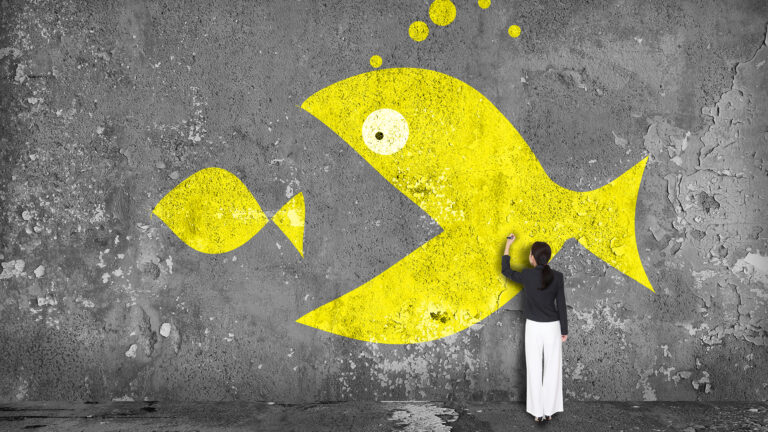 A woman paints a large yellow fish about to eat a small fish on a grey wall.