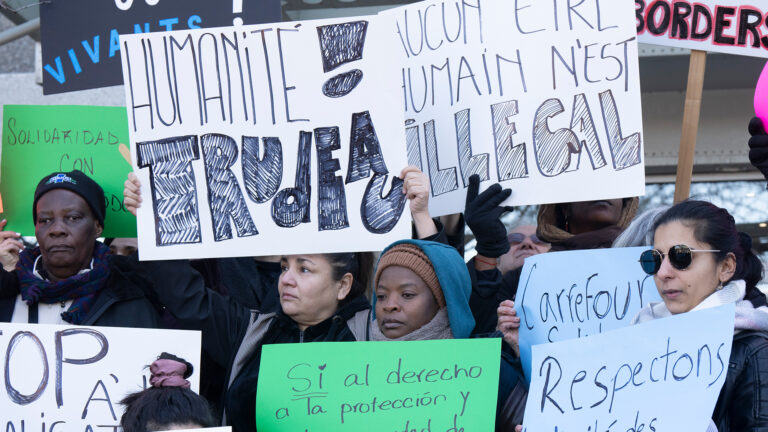 A crowd of people with four protesters in the foreground hold multiingual signs saying “no human is illegal” and “respect the dignity of asylum seekers.”