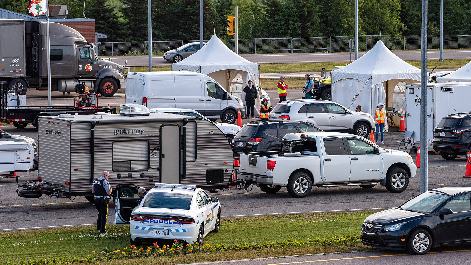 Cars, trucks and RV trailers wait in adjacent lanes to pass in front of white four-sided tents with workers wearing fluorescent yellow and orange safety vests. A police car sits nearby, with two officers.