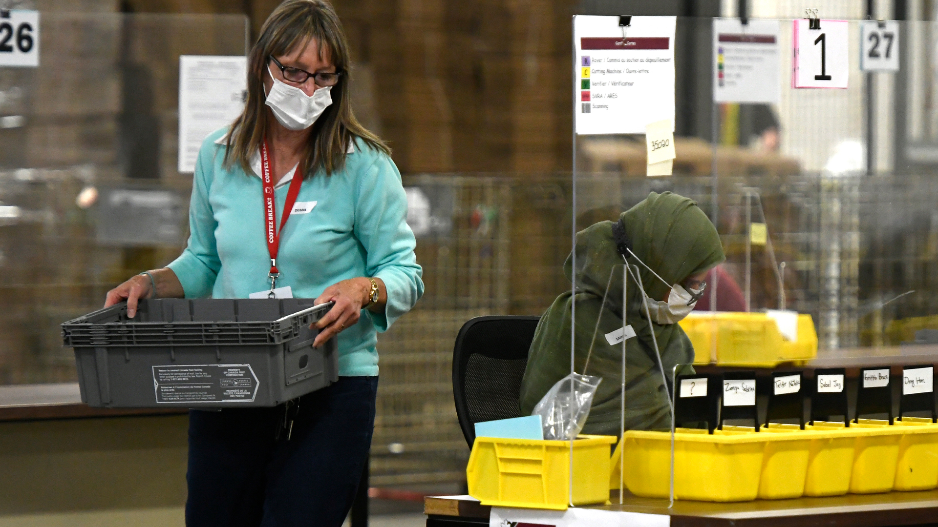A woman walks with a grey bin while another works at a table with seven smaller sorting bins in front of her.