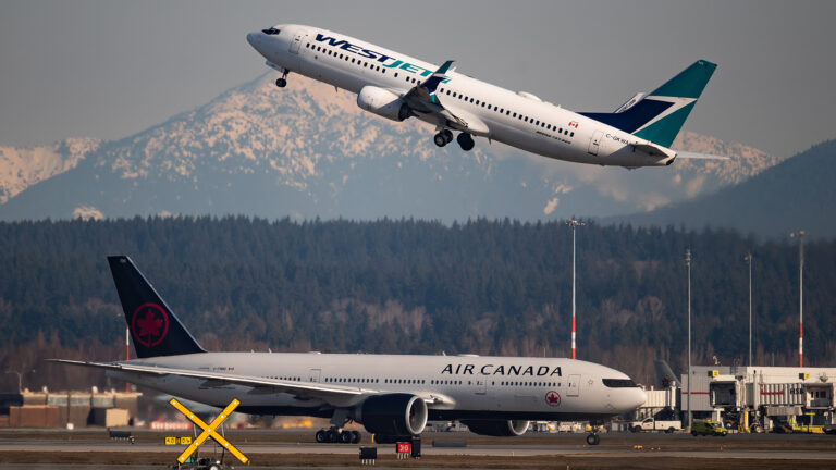 A Westjet flight soars while the Air Canada flight taxis on the ground with a snow-capped mountain in the background.
