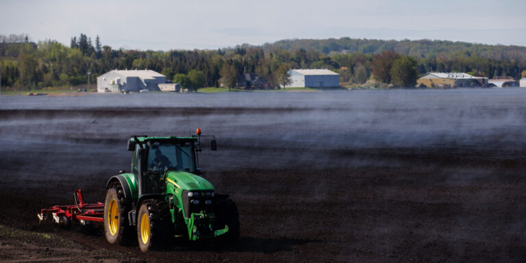ALT text: A green John Deere tractor pulls a small red tilling implement over a black field. The surface of the field is obscured by mist even though the sky is blue. Farm buildings and a forest can be seen in the background.