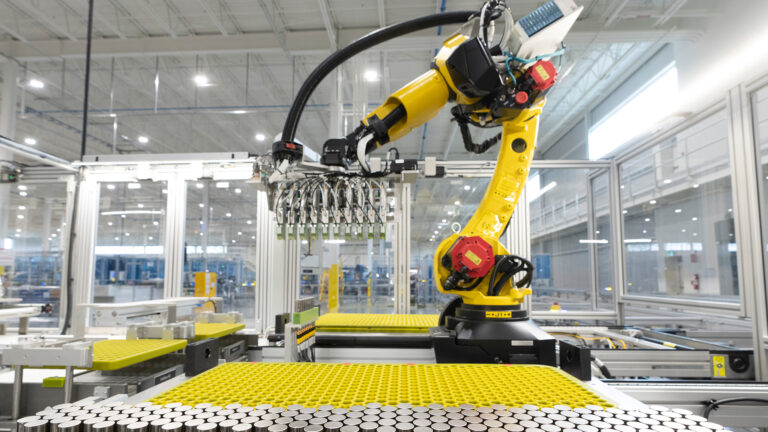 In a glass-walled room in the factory, a yellow and black robotic arm places a dozen or so small silver cylinders into a large, flat yellow surface with hundreds of holes that hold cylinders. Several dozen cylinders are already sitting in the holes.
