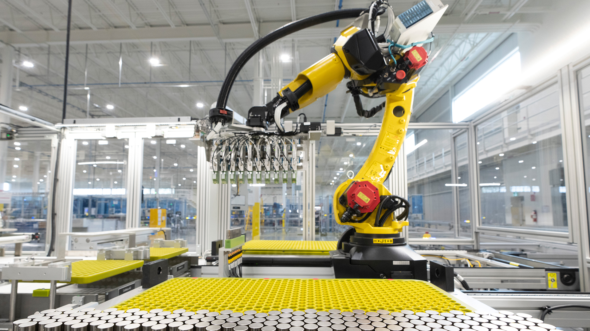 In a glass-walled room in the factory, a yellow and black robotic arm places a dozen or so small silver cylinders into a large, flat yellow surface with hundreds of holes that hold cylinders. Several dozen cylinders are already sitting in the holes.