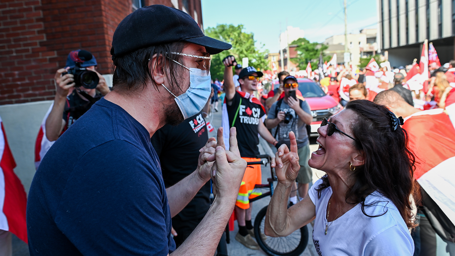 A man gives a woman the finger with both hands while she wags her finger at him. The man is wearing a mask. In the background protesters wave Canadian flags and wear T-shirts disparaging Justin Trudeau with nasty language.