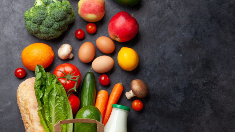 A brown paper bag filled with vegetables is laid out on a dark grey stone background. Visible are a bottle of milk, loaf of bread, lettuce, an avocado, tomatoes, cucumber, carrots, mushroom, lemon, orange, eggs, broccoli and an apple.