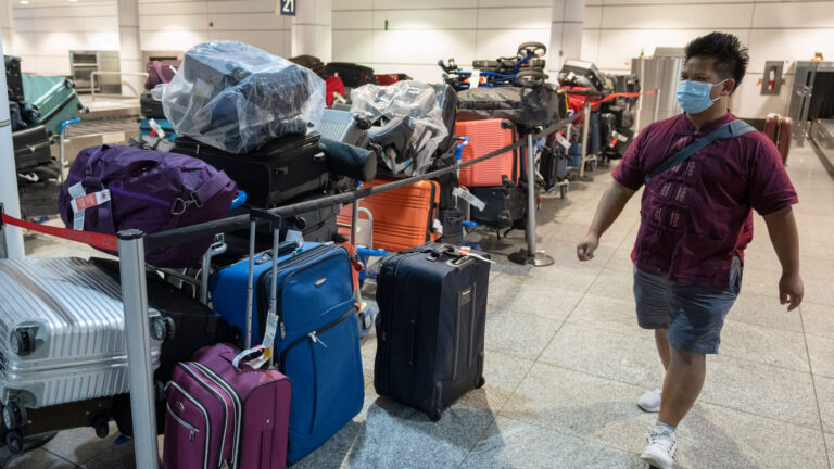 A man in t-shirt, shorts and sneakers walks past a long, cordoned-off pile of suitcases in the arrivals area of the airport.