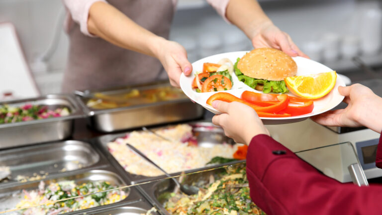 A cafeteria worker hands a plate with tomatoes, a piece of orange, and a sandwich to a child.