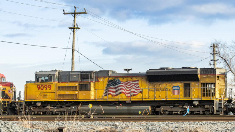 Seen from the side of a length of rail line, the locomotive is yellow with an illustration of a waving American flag painted on its side.