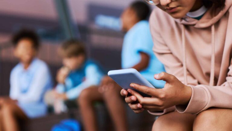 Closeup of a young girl sitting hunched over while she looks at her phone. In the background, three other kids sit together. One is laughing.
