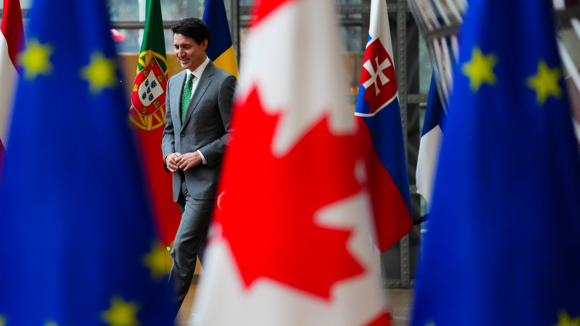 Canadian and European Union flags in foreground. In the background, ex Prime Minister Justin Trudeau walks and appears between them.