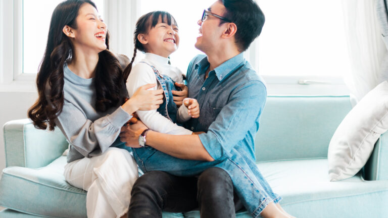 Une famille d’origine asiatique, composée d’un père, d’une mère et d’une fillette, au centre, passe un moment en riant sur un divan.