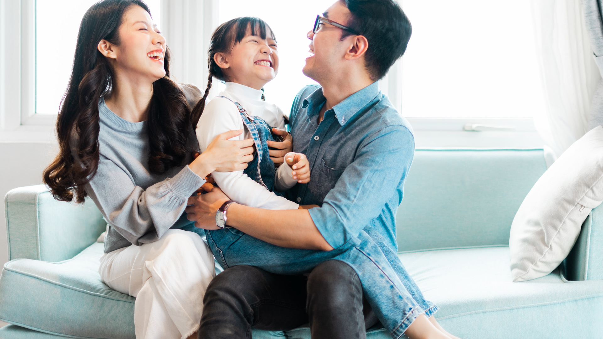 Une famille d’origine asiatique, composée d’un père, d’une mère et d’une fillette, au centre, passe un moment en riant sur un divan.
