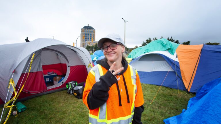 Peddle is a white woman who looks to be in her 20s. She’s wearing an oversized fluorescent orange, yellow and grey safety jacket over a black hoodie. Her ballcap is white. She’s smiling and holds two fingers in the peace sign. Behind her are several camping tents, some grey and red, some blue and turquoise. The grass is green. An elegant building sits in the distance.
