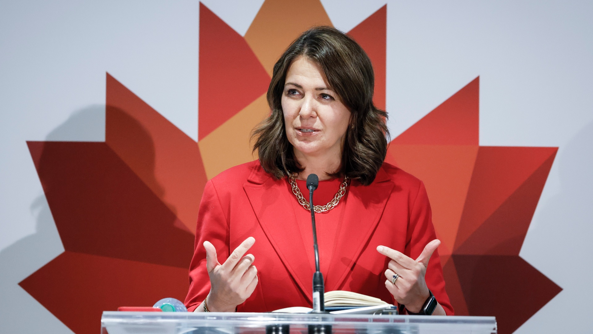 Premier Danielle Smith speaks in front of a podium wearing a red blazer and blouse stands in front of a stylized illustration of a maple leaf.