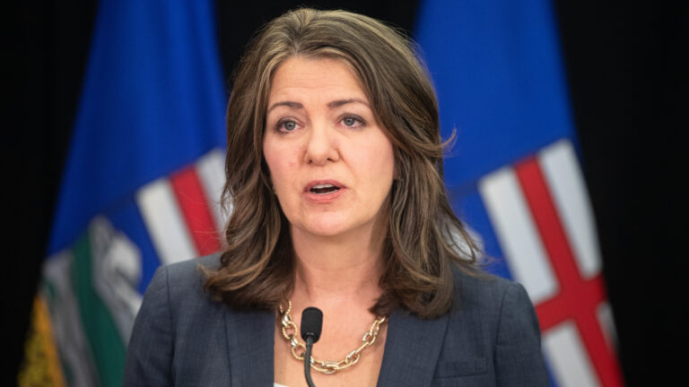 Danielle Smith speaks before microphones with two Alberta flags in the blurred background.