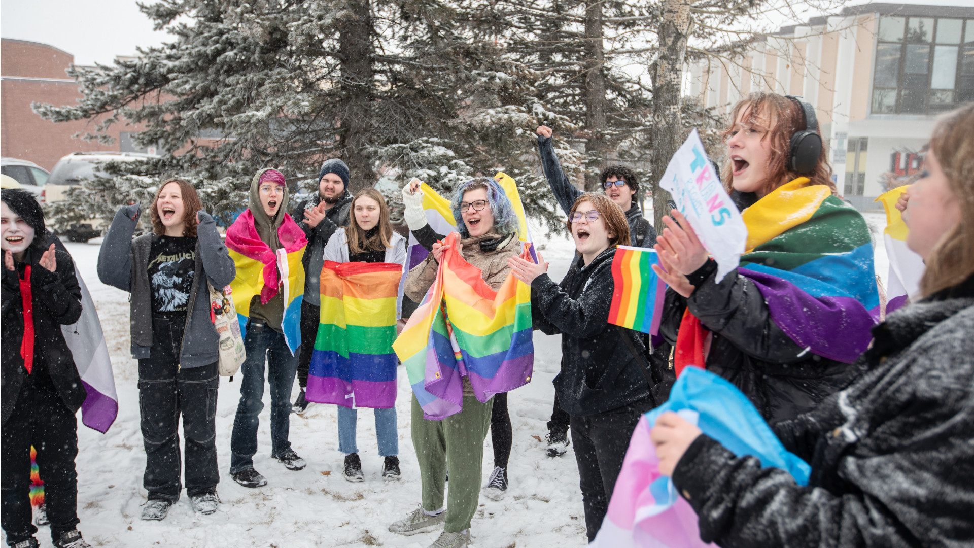 A dozen students, several wrapped in rainbow flags, stand on snowy ground while chanting.