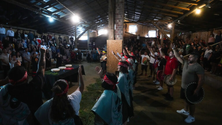 About two dozen band members raise their right arms as they stand in a line. Several wear red headbands. A small crowd stands along the walls of the large hall-like building.