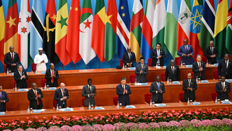 The men are standing in three rows in front of a bank of flags representing many African countries. In front of the three long wooden tables are long rows of potted flowers in light and dark pink blooms. The mean are all in dark western business suits, except one, who wears a white top, pants and flat-top white hat.