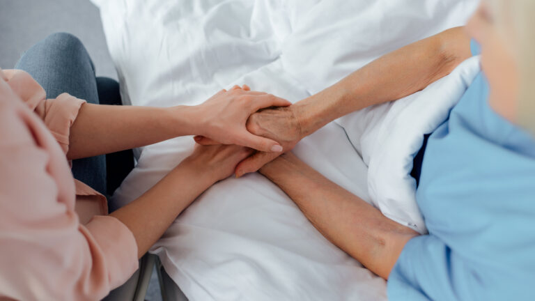 An older white woman is sitting up in a bed with white sheets. She is holding hands with aa younger woman who is in a chair next to the bed.