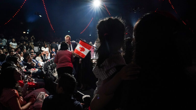 Members of a large crowd in an auditorium look on as the minister shakes hands. A small Canadian flag is in the foreground.