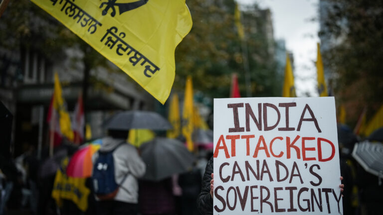A person who cannot be seen holds a sign that says “India attacked Canadian sovereignty” with protesters and yellow flags in support of the Khalistani independence movement in soft focus in the background.