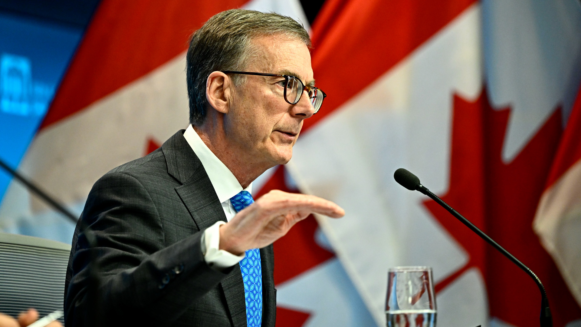 Tiff Macklem, in grey suit and blue tie, uses his hand to demonstrate a level. Two Canadian flags are out of focus in the background.