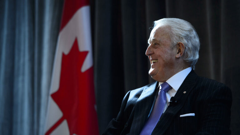 L’ancien premier ministre Brian Mulroney sourit à côté d’un drapeau canadien.