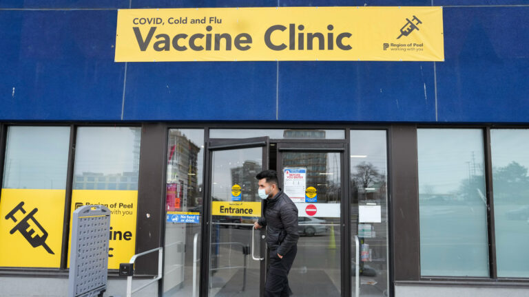 The man is holding open the door to the clinic, which has a row of tall windows framed in black. Above them a yellow sign with black letters says “COVID, Cold and Flu Vaccine Clinic, Region of Peel, working with you.” The building is a deep blue. The man is white with short black hair and a pale-blue mask over his mouth. He is wearing a black jacket, black pants and black shoes.
