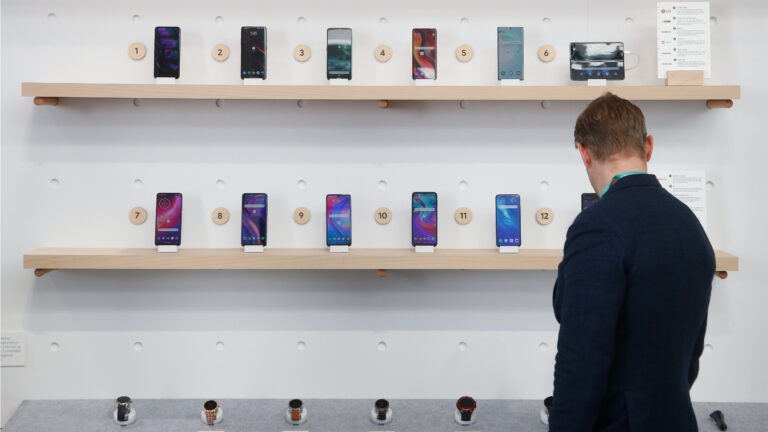 A man in a black jacket looks at a row of watches. On the wall above are two shelves of smart phones.