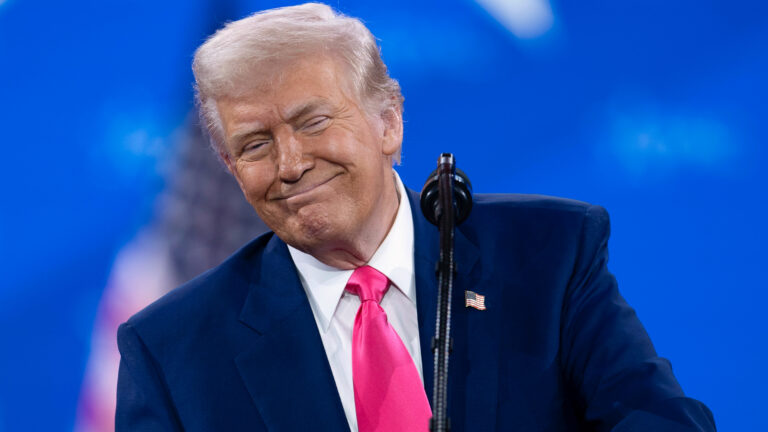 Trump is smiling widely, lips closed, eyes nearly shut, as he stands at a podium with a bright blue backdrop. He is leaning to one side of the mic. He’s in a dark blue suit, pink tie, white shirt. ¬¬