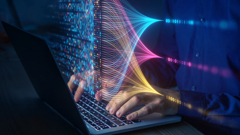 In a darkened room, a man’s hands type on a laptop. Overlayed is a translucent illustration of data in three thin rows, one blue, one magenta, one yellow, all leading into a square block of combined data that hovers over the hands and laptop.