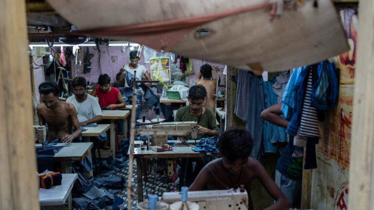 About 10 young men and boys sit at tables of sewing machines, feeding fabric into them. Piles of fabric are heaped on the floor. The walls are pink, and a dirty tarp is pinned up in an opening in the wall.
