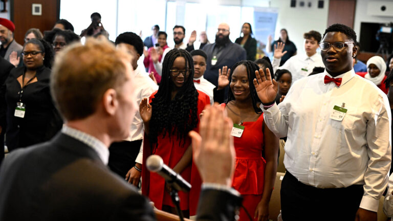 Miller has his back to the camera. A man in a red bowtie and two women in red dresses face the camera with their hands raised as they take the oath. A couple dozen other people are in the background also with a hand raised.