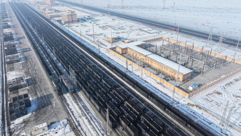 A multi-lane railroad cuts through a flat, barren and snowy landscape. About 10 tracks side by side are filled with black shipping containers filled with mounds of coal. On one side is a yard with stacked empty containers. On the other are clusters of low-rise buildings , with an empty two-lane highway running parallel.