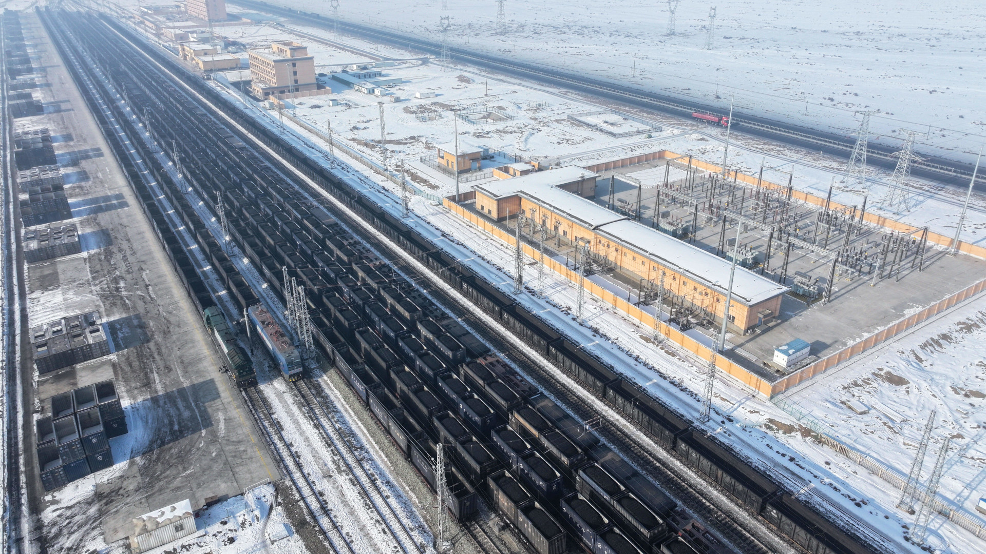 A multi-lane railroad cuts through a flat, barren and snowy landscape. About 10 tracks side by side are filled with black shipping containers filled with mounds of coal. On one side is a yard with stacked empty containers. On the other are clusters of low-rise buildings , with an empty two-lane highway running parallel.