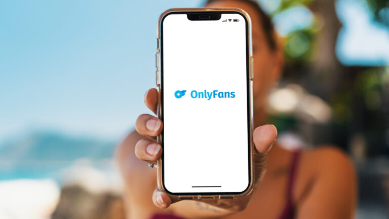 Bikini-clad girl on the beach is holding a smartphone with the OnlyFans app on the screen. Everything but the phone is in soft focus.