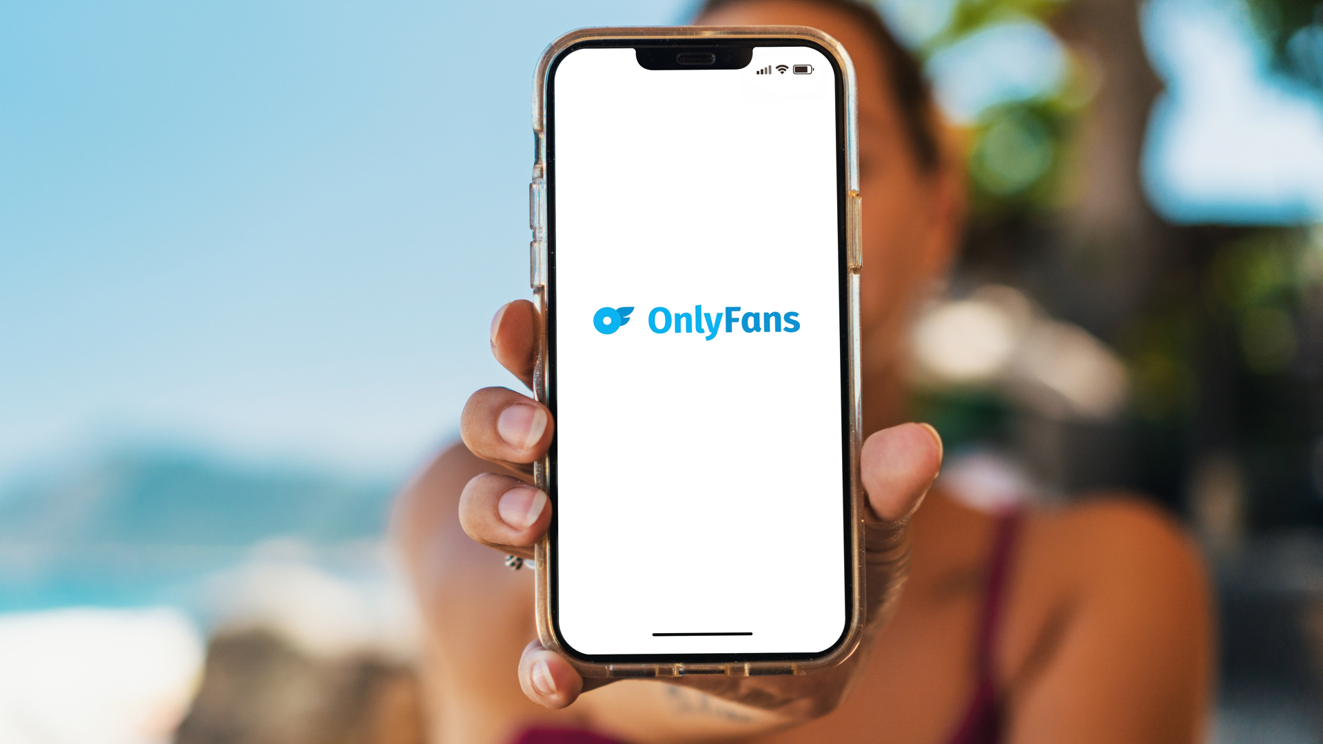 Bikini-clad girl on the beach is holding a smartphone with the OnlyFans app on the screen. Everything but the phone is in soft focus.