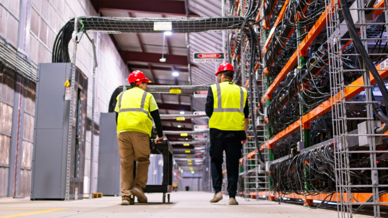 Two technicians wearing yellow high visibility coats and hard hats walking past racks of equipment in a data centre.