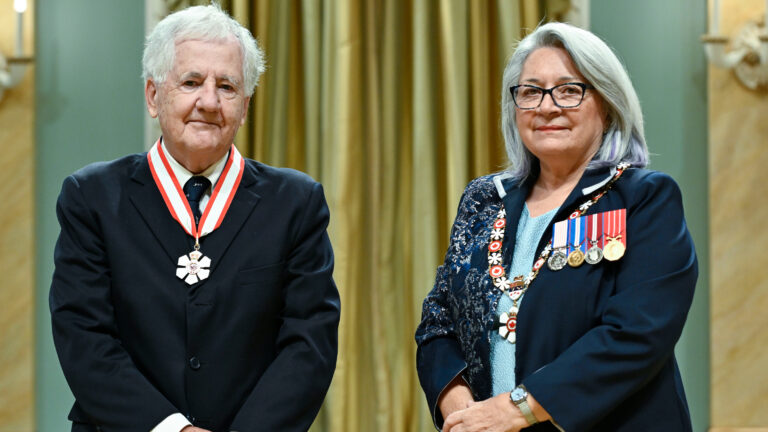Donald Savoie, in a dark suit, wears his Order of Canada medallion standing next to Mary Simon who is wearing her own medals.