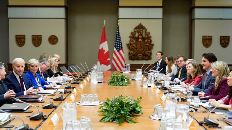 Attendees sit at a very long and very wide blond-wood table. At least 10 people sit on each side, the Americans on one side; the Canadians on the other. They each have folders, notebooks and individual microphones in front of them. The group is relaxed, with most people smiling. At the far end of the table, a Canadian and American flag hangs on tall posts. On the walls are wooden crests of arms and provincial symbols.