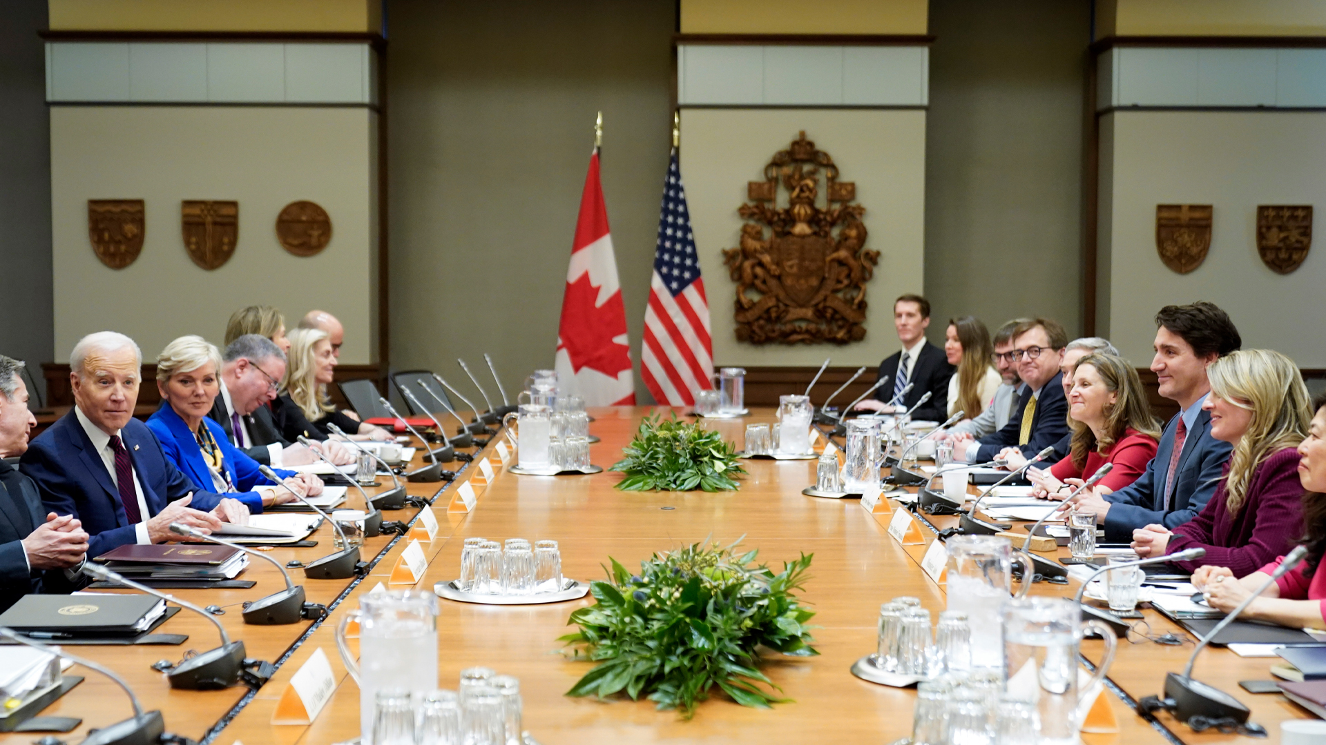 Attendees sit at a very long and very wide blond-wood table. At least 10 people sit on each side, the Americans on one side; the Canadians on the other. They each have folders, notebooks and individual microphones in front of them. The group is relaxed, with most people smiling. At the far end of the table, a Canadian and American flag hangs on tall posts. On the walls are wooden crests of arms and provincial symbols.