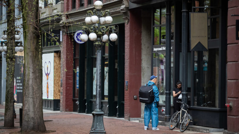 A man in a blue suit with an insulated backpack stands in front of a woman holding a plastic bag. 