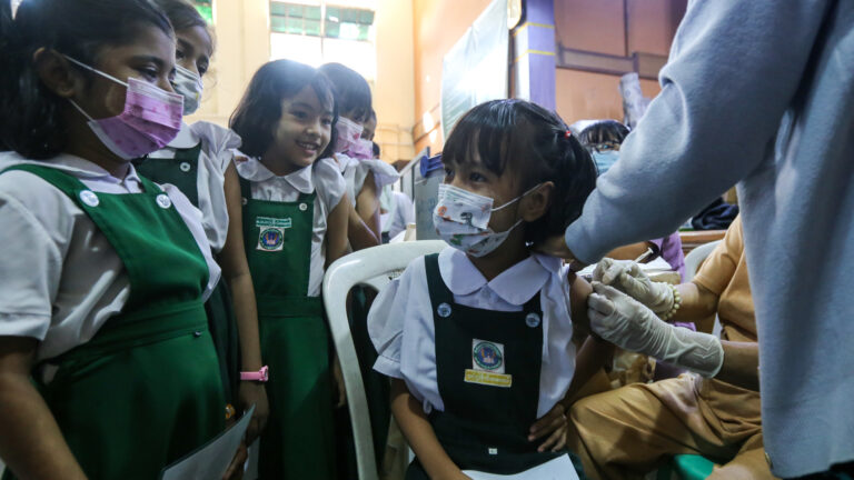 A young girl sits as she gets a needle in her arm from a woman with rubber gloves. The girl is looking away from the needle and is focusing on one of her five classmates gathered around her. They are all wearing forest-green tunics over white short-sleeved shirts, and most of them are smiling. Another adult has a hand on the girl’s shoulder.