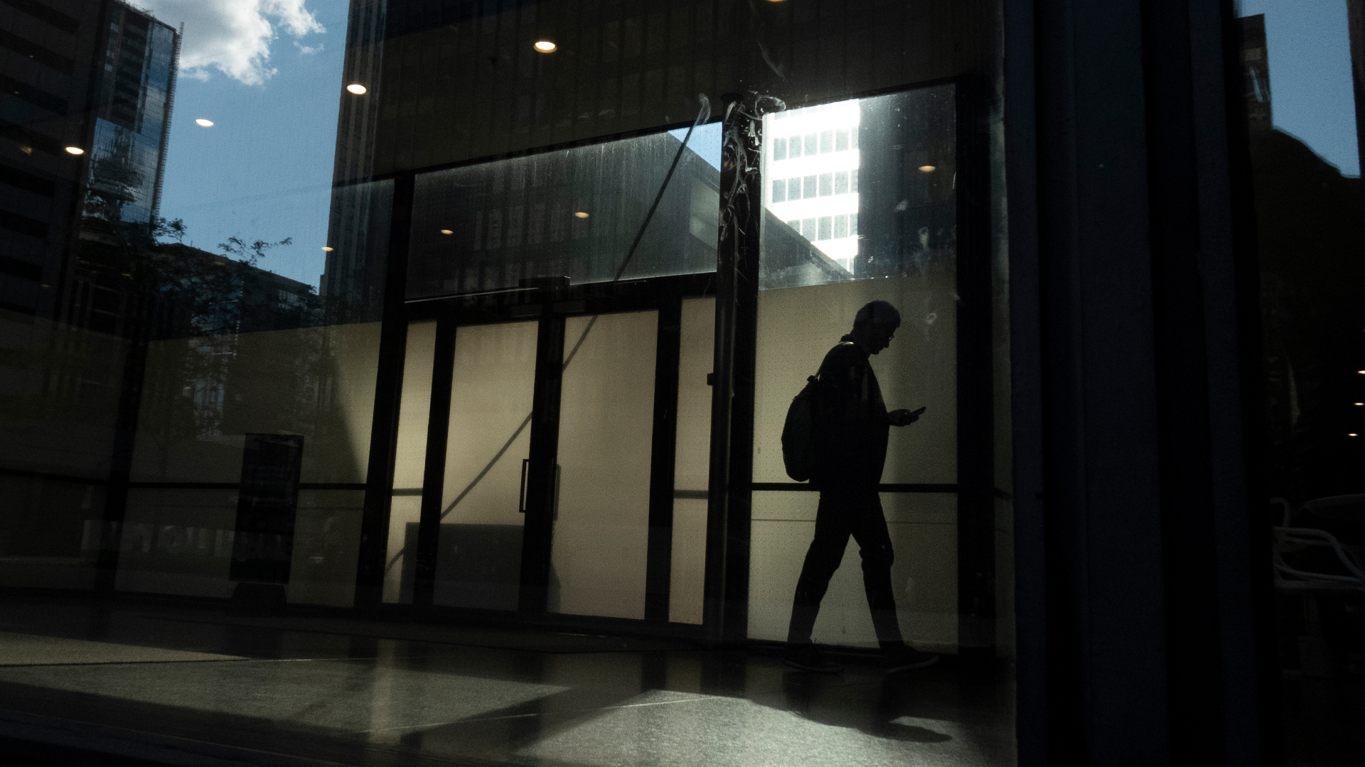 Un ouvrier en silhouette portant un sac à dos marche dans un immeuble de bureaux sombre non identifié. D’autres bâtiments et un ciel bleu ne montrant qu’un seul nuage se reflètent dans les fenêtres.